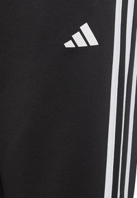 Pantalones deportivos negros con tres rayas blancas verticales y un logo blanco en el lado izquierdo, fabricados con una tela lisa y texturizada.