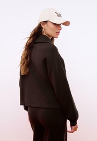 Dunkelbrauner, übergroßer Pullover mit hohem Kragen, kombiniert mit schwarzen Hosen. Eine beige Kappe mit dem Text "LA" in dunklen Buchstaben.