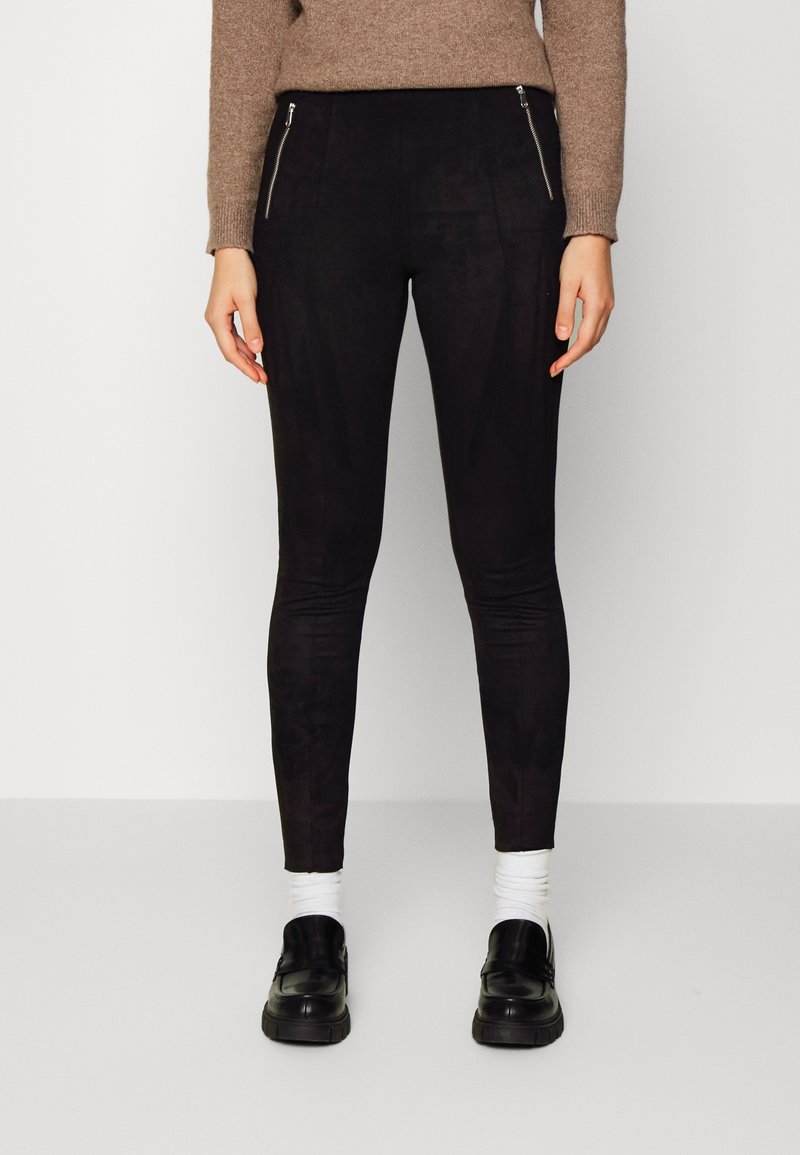 Personne portant un pantalon noir slim avec des fermetures éclair à l'avant, des chaussettes blanches, des mocassins noirs et un pull marron à manches longues, debout sur un fond uni.