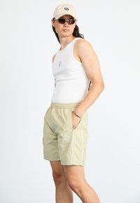 Lettvekts beige shorts med sidet piping, brodert logo, kombinert med en hvit tank-top, beige caps og grønne solbriller.