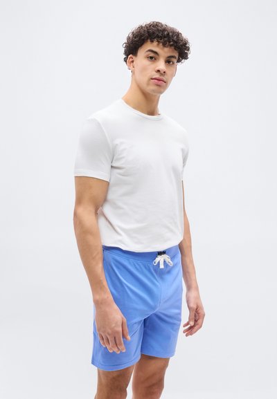 Νεαρός με κατσαρά μαλλιά που φοράει λευκό κοντομάνικο μπλουζάκι και γαλάζιο σορτς στέκεται μπροστά σε απλό λευκό φόντο.