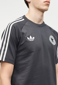 Homme portant un t-shirt Adidas gris foncé avec trois bandes blanches sur les manches, logo Adidas et emblème du Deutscher Fußball-Bund sur la poitrine.