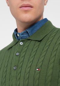 Pull en maille vert avec un col polo, comportant deux boutons noirs et un petit logo brodé avec des détails rouges, blancs et bleus.