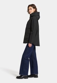 Svart vattentät jacka med hög krage, med en dragkedjestängning och två framfickor; kombinerad med utsvängda jeans i denim och svarta stövlar.