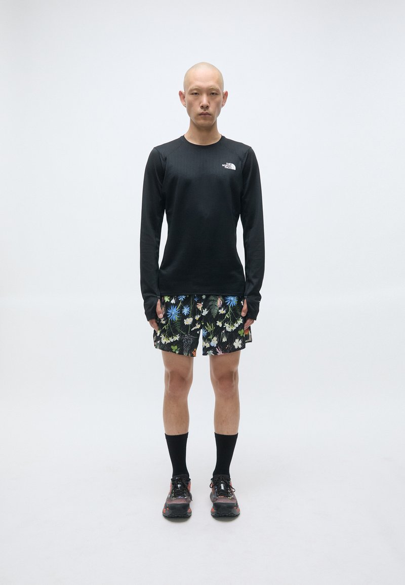 Schwarzes Langarmshirt, gemusterte Blumen-Shorts, schwarze Socken und schwarze Schuhe mit Akzenten. Glatter Stoff, sportliche Passform.