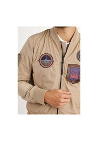 Veste bomber beige avec fermeture éclair, poignets côtelés et patches brodés présentant divers motifs sur la poitrine et les manches.