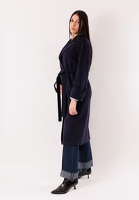 Donna in piedi con un cappotto blu navy con cintura, jeans a gamba larga con risvolto e stivaletti neri con tacco, su uno sfondo chiaro e semplice.