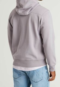Light gray hoodie with a hood and ribbed cuffs, featuring a soft texture and visible stitching. Worn with a layered white shirt beneath.