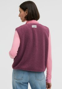 Fleece-Weste in tiefem Lila mit einer weichen, strukturierten Oberfläche, ausgestattet mit einem Etikett auf der Rückseite. Getragen über einem hellrosa Langarmoberteil.