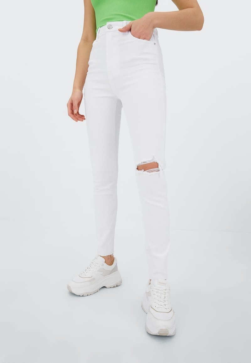 Jean blanc taille haute avec un genou droit déchiré, associé à un haut moulant vert et des baskets blanches épaisses. Texture lisse, coupe slim.