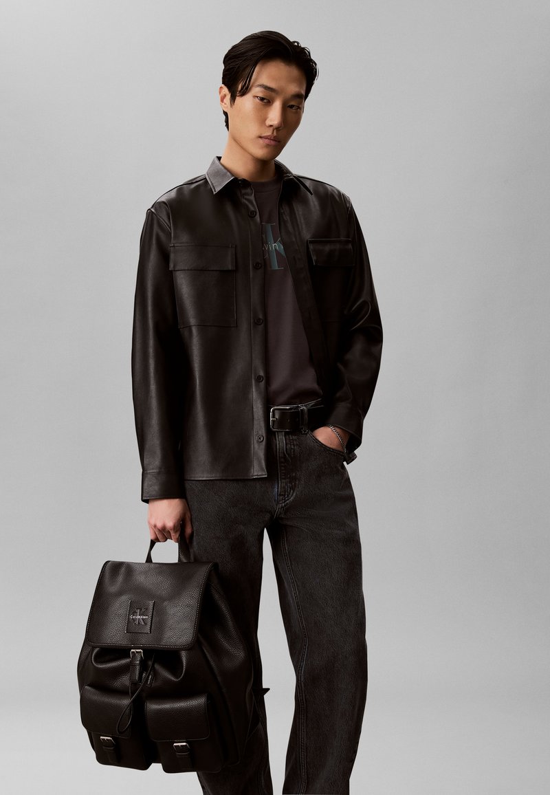 Jeune homme portant une chemise en cuir noir et un jean foncé, tenant un sac à dos en cuir noir dans une main devant un fond gris uni.