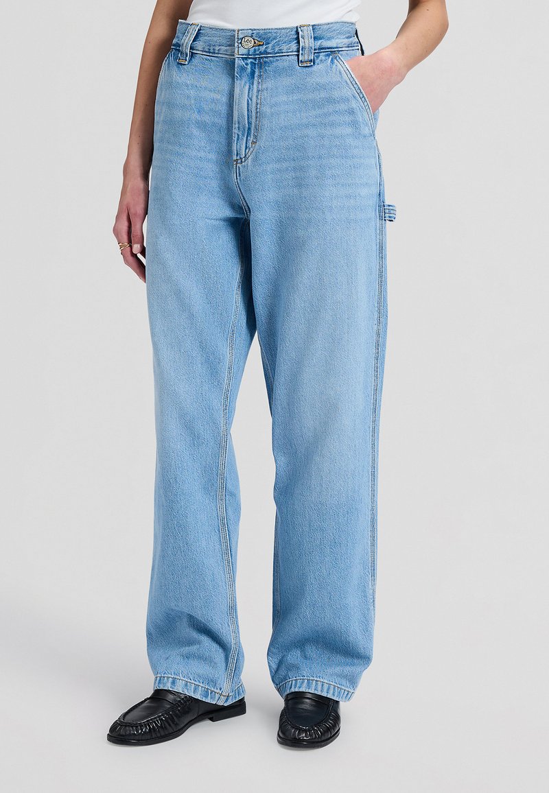Jean bleu clair coupe large avec boucle latérale, porté avec des mocassins noirs et un haut blanc, main reposant dans la poche.