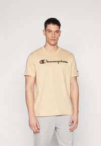 Beige Baumwoll-T-Shirt mit Rundhalsausschnitt und kurzen Ärmeln. Mit einem schwarzen "Champion"-Logo auf der Vorderseite, mit einem roten Akzent im "C."