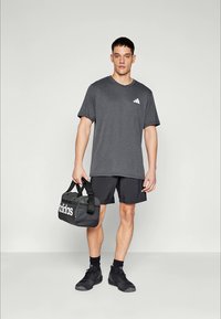 Graues sportliches T-Shirt mit kurzen Ärmeln, versehen mit einem weißen Logo. Schwarze Shorts und eine schwarze Sporttasche mit einem weißen Logo, kombiniert mit schwarzen Turnschuhen.