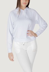 Felpa bianca con cappuccio, dal fit rilassato e con cordini regolabili; abbinata a pantaloni bianchi aderenti con un logo in dettagli argentati.