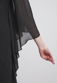 Bras droit tendu vers le bas, portant une longue manche noire fluide et transparente sur un vêtement noir, sur un fond clair uni.