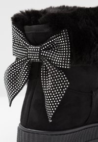 Svart mocka känga med fuskpälsdetalj, med en stor svart och silvern studsad rosett vid vristen. Texturerad sula och vadderad krage.