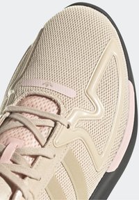 Beige athletic shoe with mesh upper, pink accents, plastic overlays, and grey laces. Features padded collar and textured rubber sole.