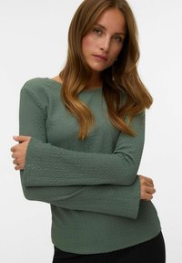 Groene textuur top met een afgeronde halslijn en lange klokmouwen. De stof heeft een subtiel kreukpatroon. Draagt over donkere kleding.