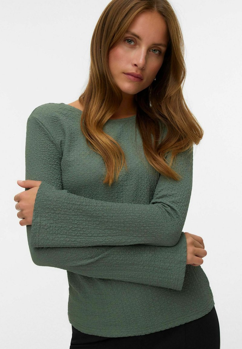 Groene textuur top met een afgeronde halslijn en lange klokmouwen. De stof heeft een subtiel kreukpatroon. Draagt over donkere kleding.