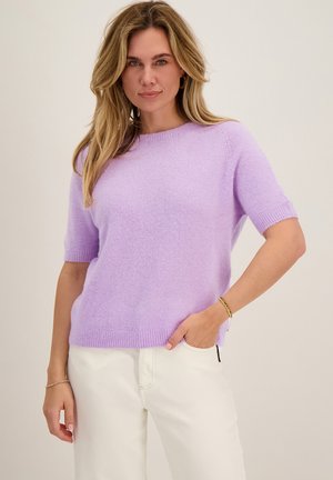 Vrouw met lang blond haar die een lavendelkleurige trui met korte mouwen en witte broek draagt, staand met één hand in de zak tegen een effen achtergrond.