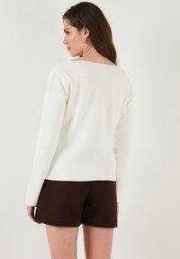 White, long-sleeve sweater with a textured weave, featuring a low back neckline. Paired with brown shorts, showcasing a relaxed fit.