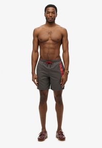 Grijze zwembroek met rode trekkoord en logo, gemaakt van lichtgewicht materiaal. Het model is blootvoets en draagt donkerbruine schoenen.