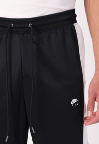 Črne športne hlače z elastičnim pasom, vrvico in belimi stranskimi paneli. Na nogi imajo črn kvadratni Nike Air logo.