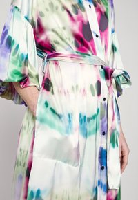 Robe-chemise tie-dye colorée avec manches bouffantes, boutons noirs sur le devant, ceinture à la taille et mains posées décontractées dans les poches latérales.