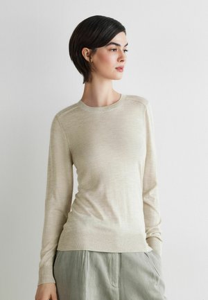 Vrouw met kort donker haar die een beige trui met lange mouwen en lichtgrijze broek draagt, staand tegen een witte muur, handen in de zakken.
