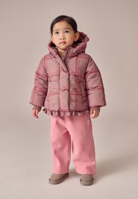 Giacca a vento rosa chiaro con orlo arricciato e motivo a quadri, abbinata a pantaloni della tuta rosa coordinati e stivali grigi. Texture liscia su tutta la superficie.