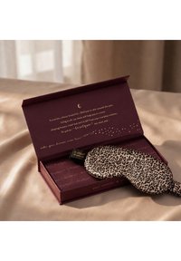 Maschera per gli occhi in seta con stampa leopardata appoggiata su una scatola bordeaux aperta con testo dorato, posta su una superficie in tessuto beige morbido.