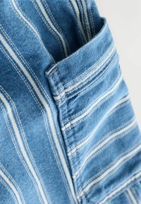 Țesătură din denim albastru cu dungi verticale albe, cu un detaliu de buzunar. Textura este netedă, cu accente de cusătură vizibile.