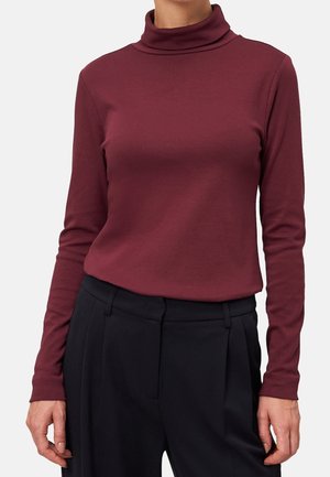 Woman wearing a long-sleeve maroon turtleneck tucked into high-waisted black pleated trousers, standing against a plain background.