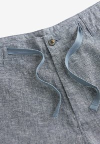 Gros plan sur la ceinture d’un pantalon bleu en chambray avec un bouton marron, des passants de ceinture et un cordon bleu, mettant en évidence les détails des coutures.
