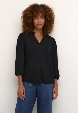 Blouse noire avec un décolleté en V, un col volanté et des manches trois-quarts. Le tissu a une finition texturée. Associé à un jean bleu.