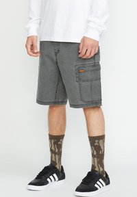 Graue Cargo-Shorts mit Seitentaschen, kontrastierenden Nähten und einem Volcom-Logopatch. Tragen Sie dazu gemusterte braune Socken und schwarze Sneaker.