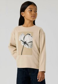 Beżowa koszulka z długim rękawem z botaniczną ilustracją kwiatu w czerni i bieli, umieszczoną na jasnobrązowym tle. Lekko luźny krój.