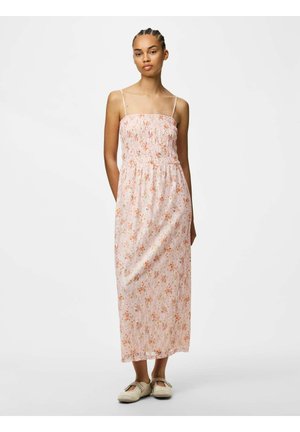 Žena s pletenými vlasmi, oblečená v ružových kvetovaných šatách bez rukávov až po členky a béžových plochých topánkach, stojaca s rukami za chrbtom.