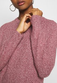 Maglione lavorato a maglia in una tonalità di rosso-rosa, con scollo rotondo e maniche lunghe con polsini a coste. Texture morbida, materiale caldo.