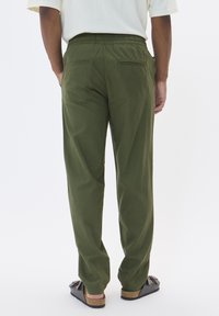 Grüne, lässige Hose mit elastischem Hüftbund, ausgestattet mit zwei Gesäßtaschen und einem weichen, strukturierten Stoff. Getragen mit dunklen Sandalen.