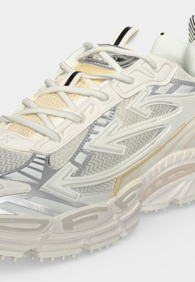 Gros plan d'une chaussure de sport blanche et argentée en tissu mesh, motifs fléchés, semelle épaisse et accents beiges.