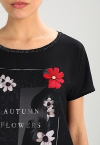 Camiseta negra con escote redondo adornado con cuentas, estampados florales en rojo y blanco, y el texto "FLORES DE OTOÑO" en plata.