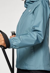 Vattentålig teal-jacka med huva; har dragkedjestängning och elastiska ärmslut; logotyp på bröstet; håller vandringsstavar.
