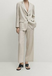 Costume en lin beige clair avec une veste sur mesure et un pantalon à jambes larges. Des sandales à talons noires à bout ouvert complètent la tenue.