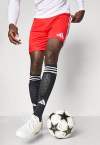 Röda träningsshorts med vitt Adidas-logotyp, parat med svarta knästrumpor som har texturerad dämpning och vita ränder. Fotboll vid foten.
