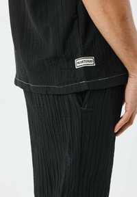 Zwart textuur tweedelig outfit met verticale streepjes, met een label aan de zoom van het shirt en een zakdetail op de broek.