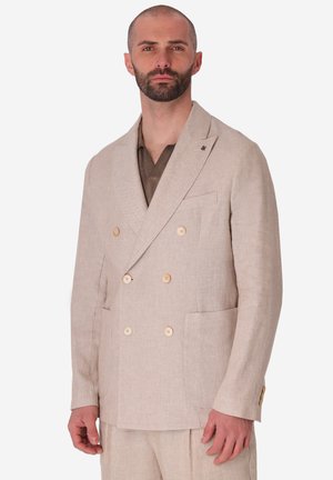 Uomo con barba corta che indossa una giacca beige doppiopetto e pantaloni abbinati sopra una camicia marrone, in piedi contro uno sfondo neutro.