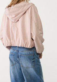 Jasnoróżowa, krótka kurtka z kapturem, wykonana z miękkiego, gładkiego materiału. Posiada elastyczny dół i obniżone ramiona, zestawiona z niebieskimi dżinsami.