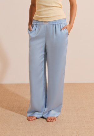 Personne portant un pantalon large en satin bleu clair et un débardeur beige, debout pieds nus sur un sol texturé.